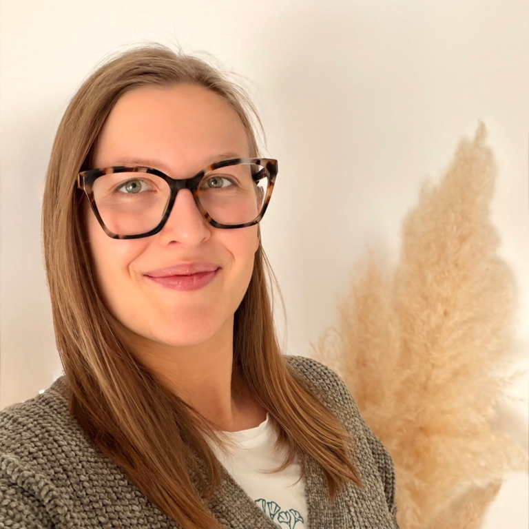 Privates Foto von Lara Katharina Aberle. Frau mit Brille und langem Haar, lächelnd vor einem hellen Hintergrund mit trockenem Gras.