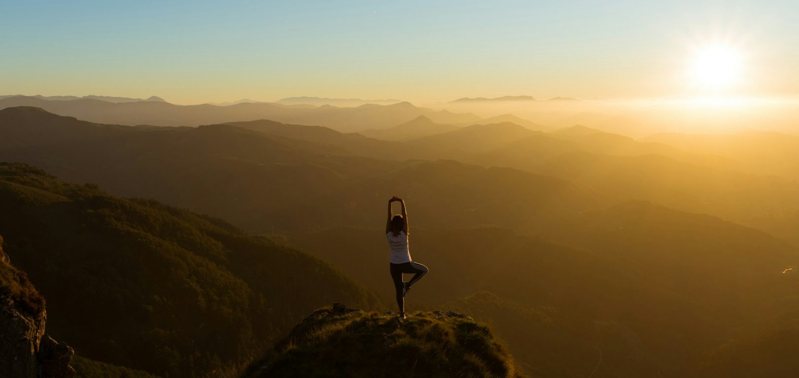 Foto von Eneko Uruñuela von Unsplash Person steht in Yoga-Pose auf einem Felsen mit Sonnenuntergang im Hintergrund.