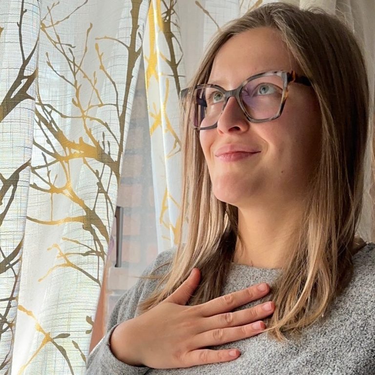 Privates Foto von Lara Katharina Aberle. Junge Frau mit Brille steht vor einem Fenster und legt ihre Hand auf das Herz.